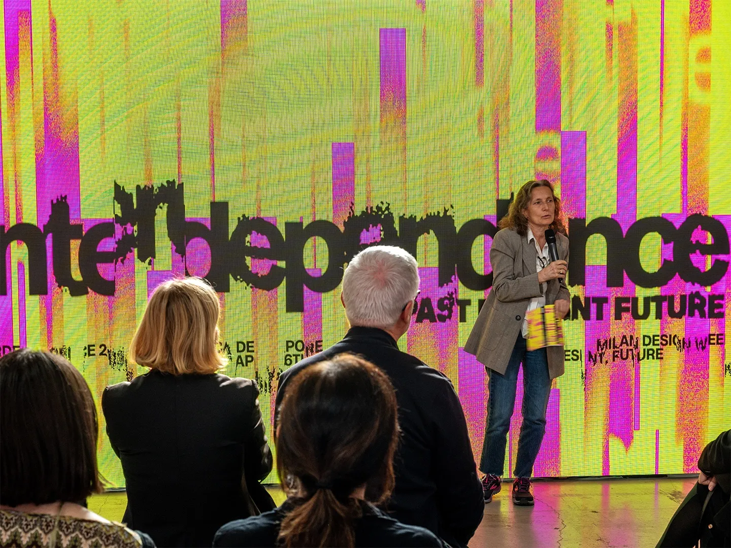 A speaker in front of a screen showing the INTERDEPENDENCE identity