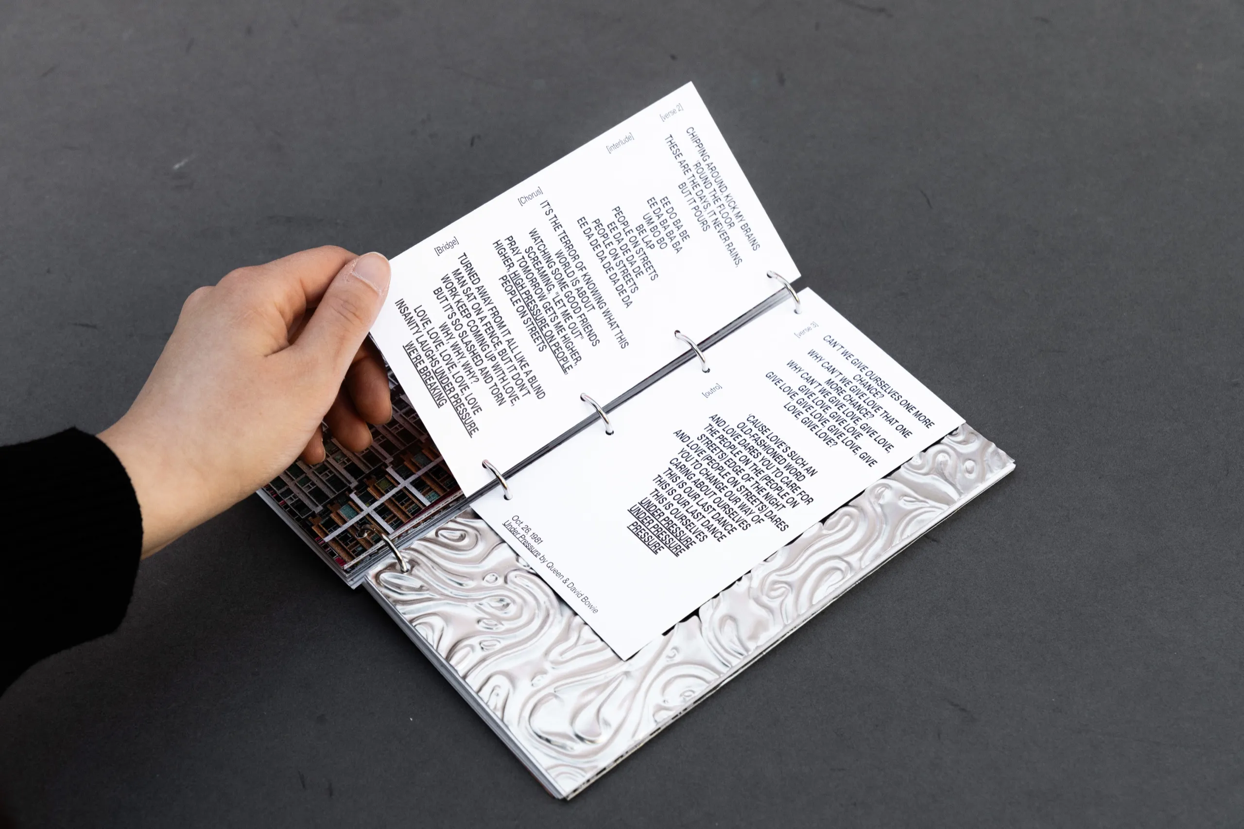 A hand turning the pages of Hostilia, above an embossed metal sheet