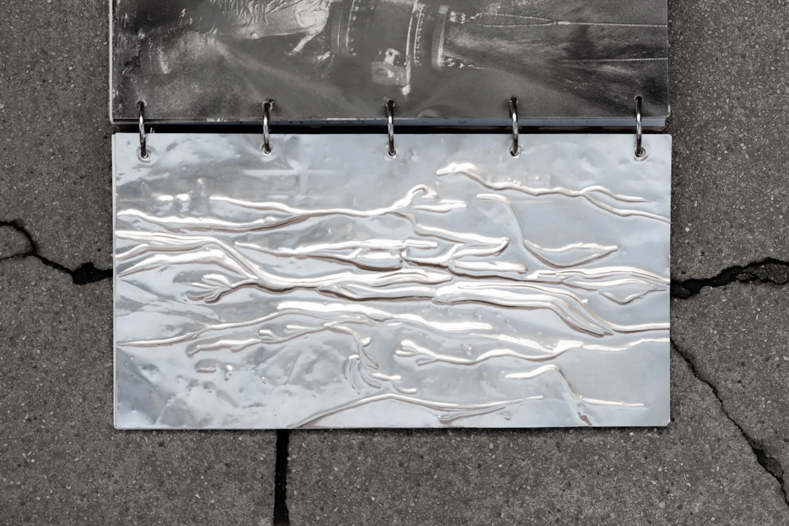 An embossed aluminium sheet bound with rings into Hostilia, on cracked concrete